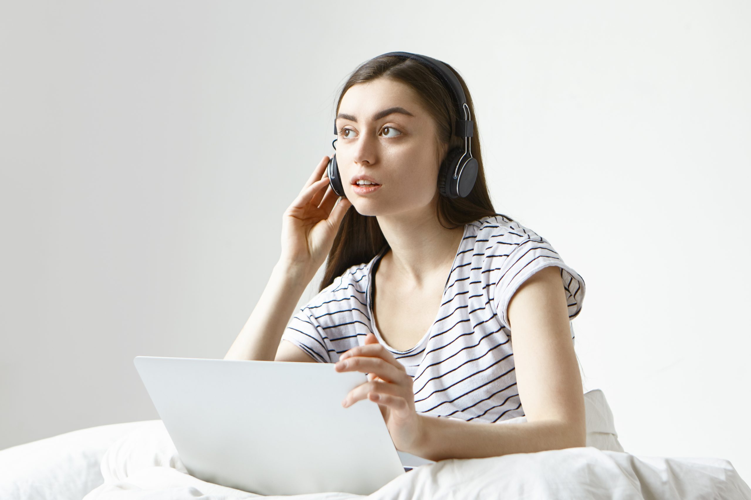 Cute young female translator working remotely from home, sitting on white bed with portable computer, listening attentively to audio file using wireless headphones, having focused serious look